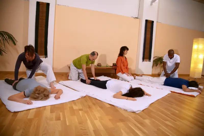 In un'ampia sala con pavimento in legno, diversi allievi di una scuola di shiatsu si esercitano in coppia su materassini (futon) bianchi. L'immagine cattura un momento di pratica in cui alcuni studenti, in ginocchio, applicano tecniche di pressione e manipolazione sulla schiena e sulle gambe dei compagni, che sono sdraiati proni. La disposizione delle persone e la presenza di più postazioni di lavoro simultanee indicano un ambiente di apprendimento guidato, tipico di una classe o di un corso di 