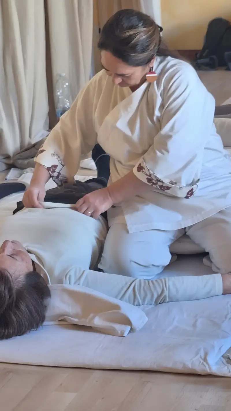 Un'operatrice shiatsu, che indossa una casacca bianca da lavoro (samue), è inginocchiata su un futon e si china su una persona sdraiata a pancia in su. L'operatrice ha le mani sul torace del ricevente, in una posa che indica l'applicazione di una tecnica o una fase di ascolto del corpo. La persona che riceve il trattamento ha gli occhi chiusi e appare in uno stato di abbandono e rilassamento.  La scena trasmette un senso di cura, concentrazione e professionalità, tipico di una seduta individuale